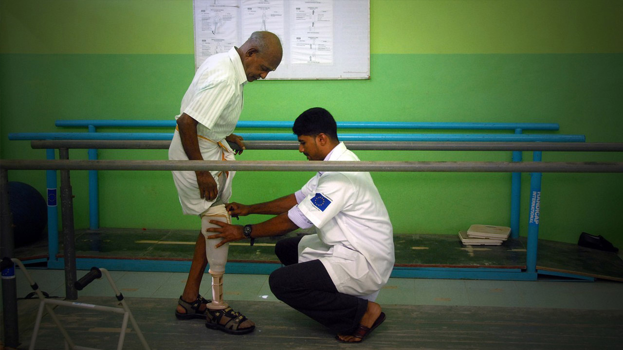 Ein Mann steht zwischen zwei Stangen zur Gehhilfe und ihm wird eine Prothese angepasst - HI Sri Lanka