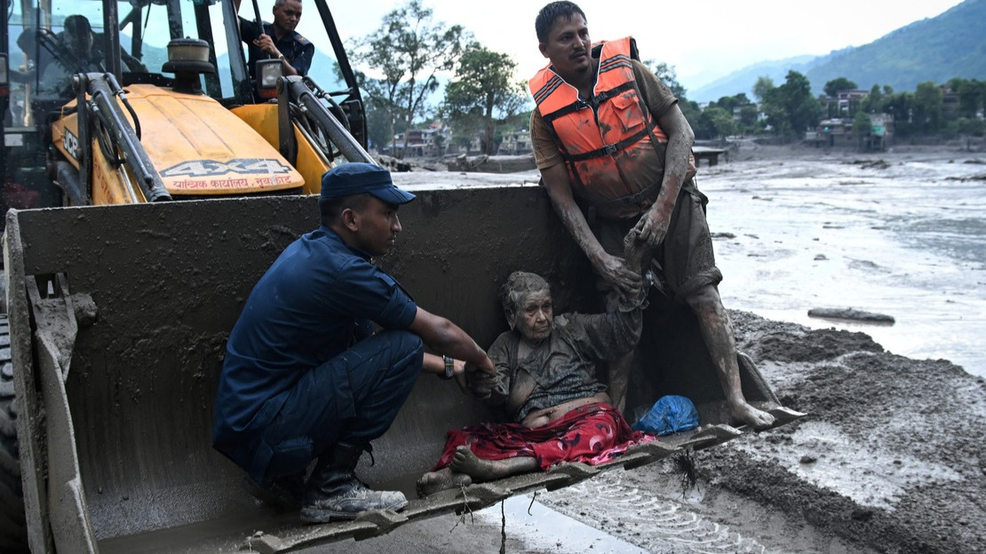 Deux hommes soutiennent une femme handicapée a demi allongée dans la benne d'un tractopelle. Tous sont couverts de boue, le paysage autour d'eux est inondé.
