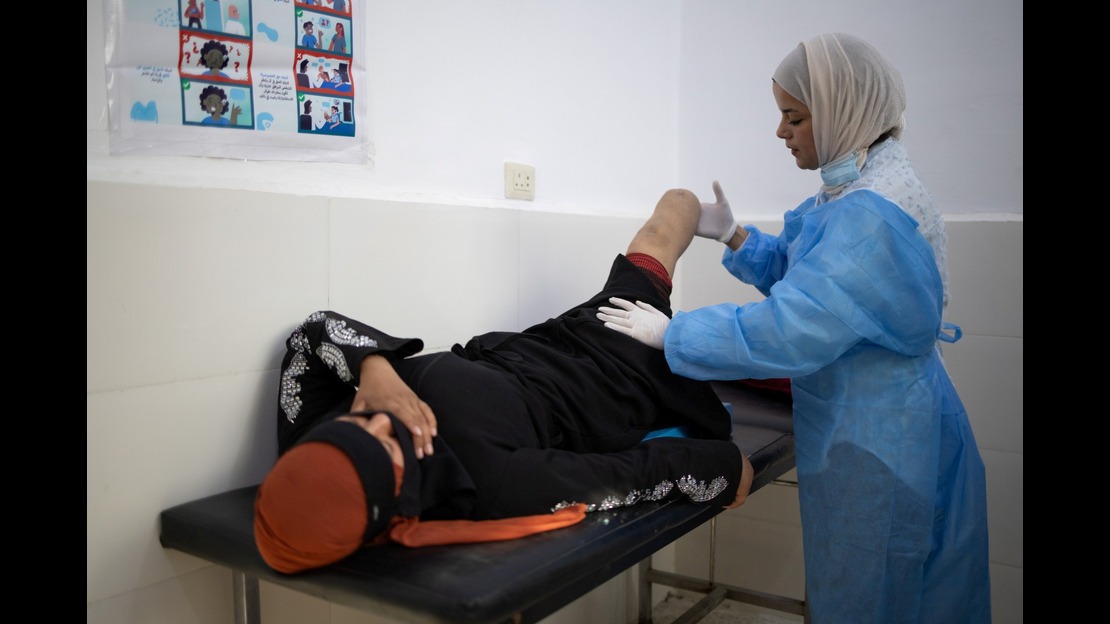 Une personne est allongée sur une table d’examen dans une pièce médicale. Cette personne est amputée d’une jambe et le moignon est visible. La personne porte des vêtements sombres avec des motifs brillants sur les manches, ainsi qu’un tissu rouge couvrant la tête. Elle est allongée sur le dos, avec une main posée près du visage. À côté de la table, une autre personne porte une tenue médicale composée d’une blouse bleue, de gants et d’un masque placé sous le menton. Cette personne manipule le moignon avec ses mains. La pièce comporte des murs blancs .