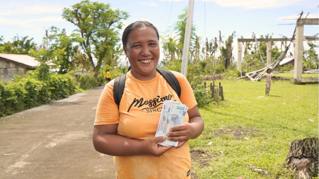 Une femme sourit à la caméra et tient quelques billets de banque. À l'arrière-plan, on aperçoit des poteaux électriques renversés.