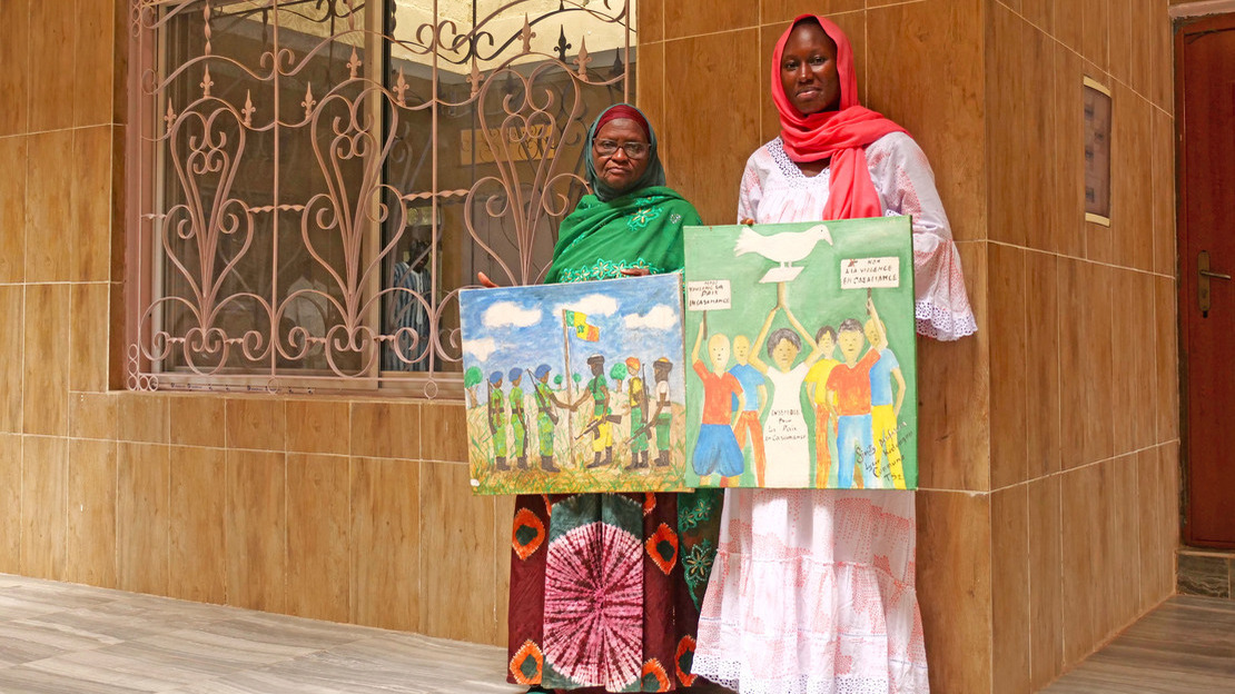 Deux femmes se tiennent debout devant un bâtiment et tiennent à la main des tableaux colorés figurant des combattants qui se serrent la main et des enfants avec des pancartes et une colombe blanche.