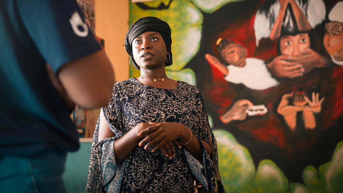 Awa est debout et regarde une personne qui se tient devant elle et dont on devine la manche du t-shirt sur lequel figure un logo HI. Derrière elle, un mur avec des peintures figurant une jeune fille qui se cache le visage avec les mains, une autre qui est enceinte, une troisième qui pleure tandis que deux mains l'empêchent de parler.