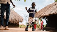 En Ouganda, un jeune garçon de 9 ans se tient debout et marche seul grâce à deux orthèses. Il sourit en regardant le photographe