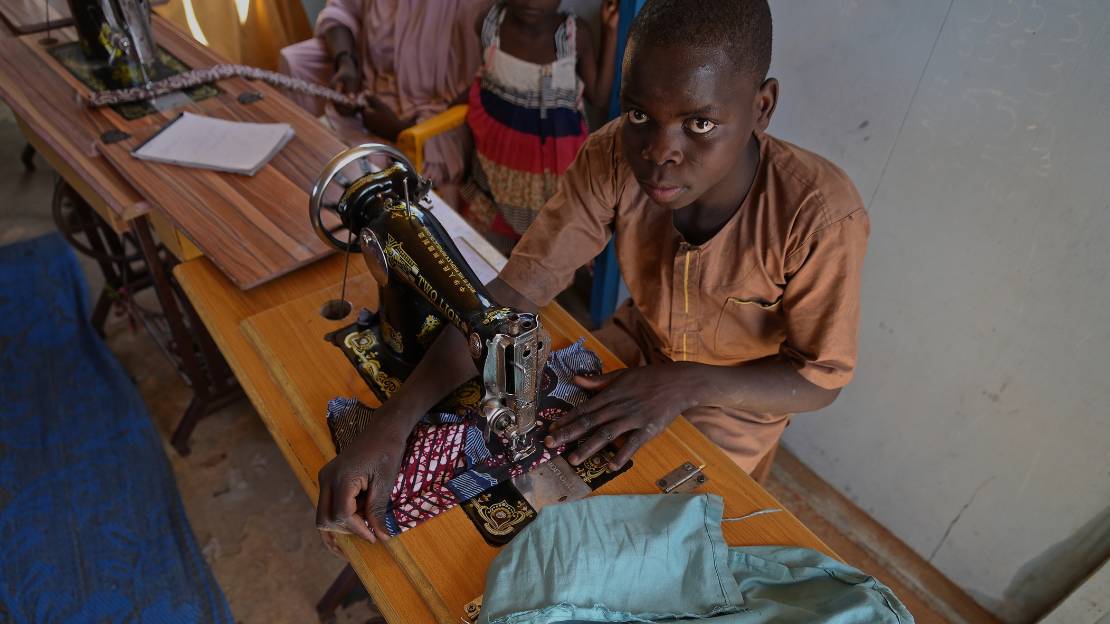 Un jeune homme est assis et utilise une machine à coudre tout en regardant l'objectif de l'appareil photo. 