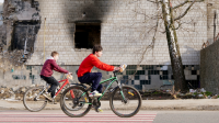 Deux garçons pédalent sur leur vélo en passant devant un bâtiment bombardé.