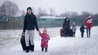 Une femme tient la main d'un enfant marchant dans la neige. à l'arrière plan, une famille avec des valises.